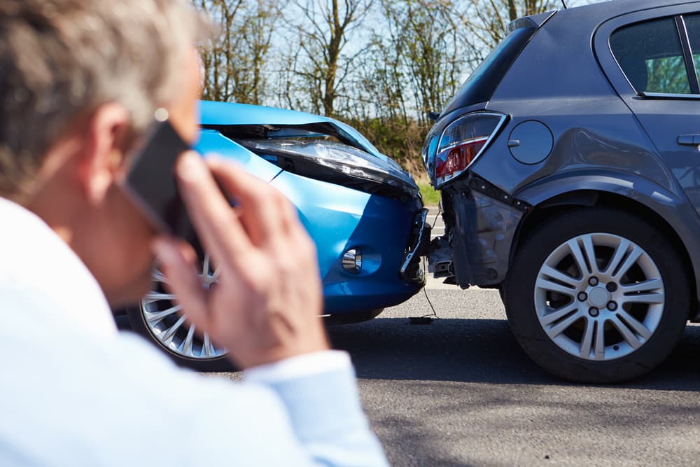 Driver on phone after rear-end car accident between blue and gray vehicles on a sunny day, showing visible bumper damage.